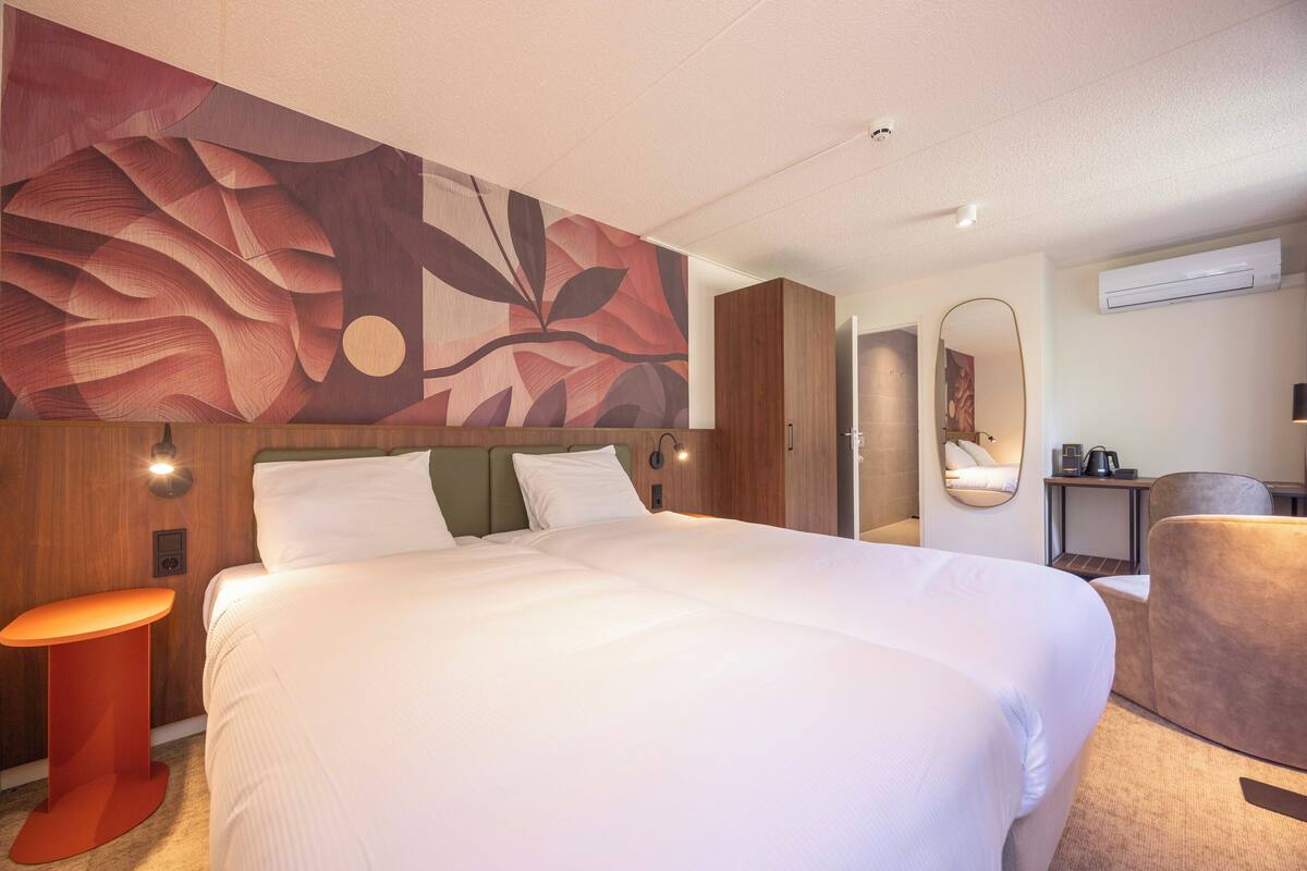 A hotel room with a large bed, a round side table, a mirror, and a wall-mounted air conditioner.