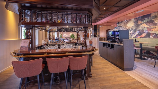 A bar area with a wooden bar counter, multiple stools, and a variety of bottles and glasses. - Fletcher Hotel - Restaurant Victoria - Hoenderloo (Hoenderloo)