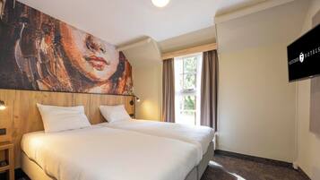 Ein Hotelzimmer mit einem großen Bett, einem Holzkopfende, einem Bild an der Wand, einem Fernseher und einem Fenster mit Vorhängen.