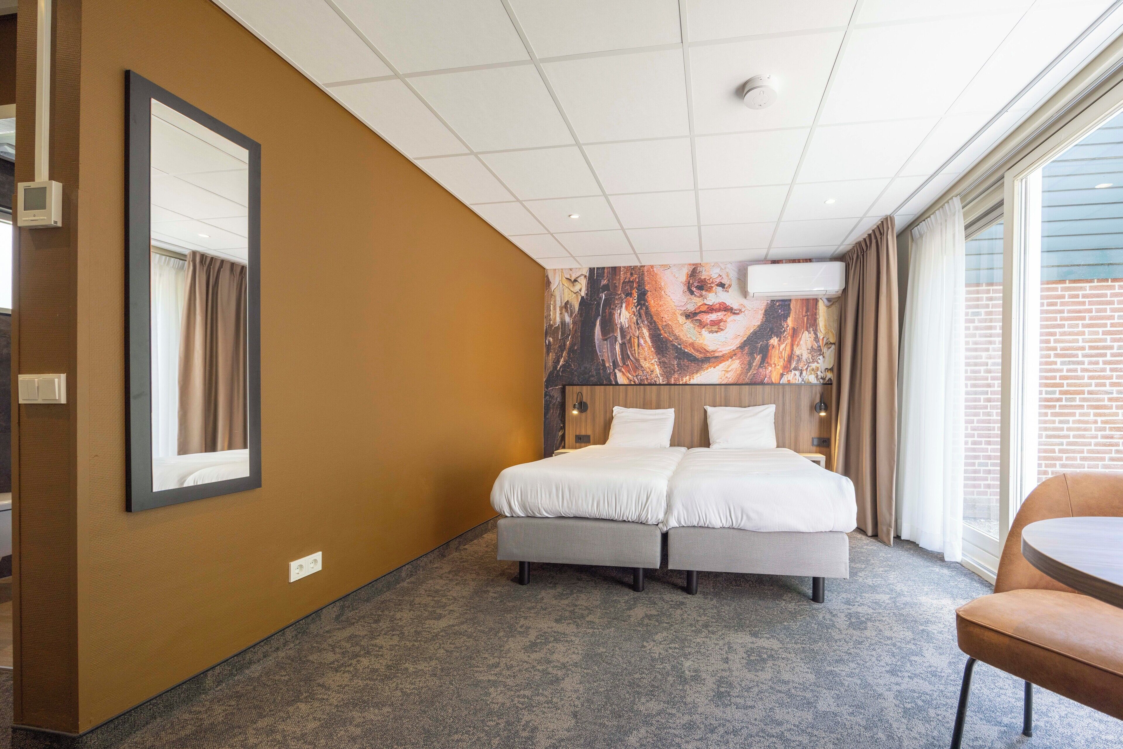 Een hotelkamer met een tweepersoonsbed, een grote spiegel, een schilderij aan de muur en uitzicht op het omliggende landschap.