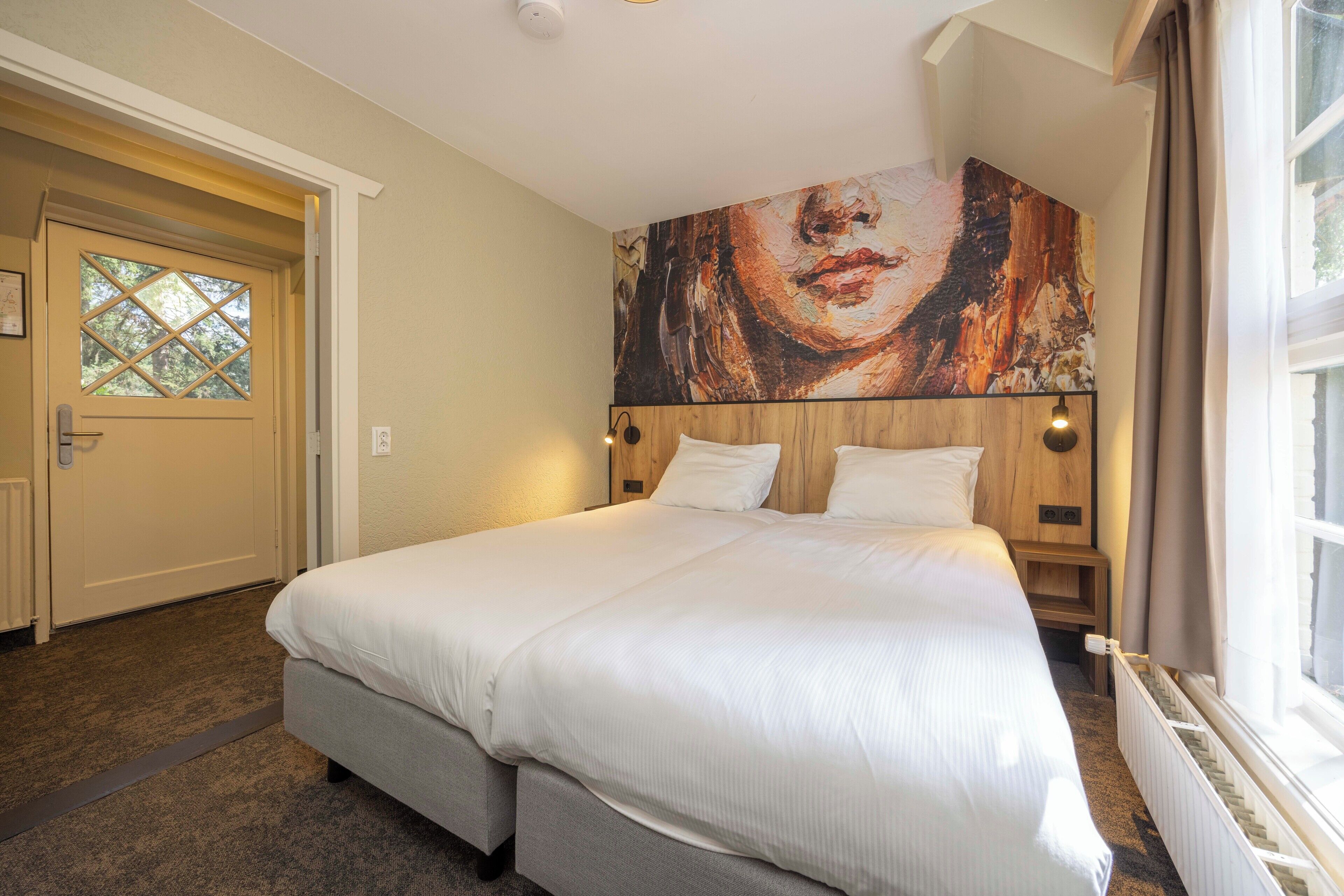 A hotel room with a large bed, a wooden headboard, a window with curtains, and a wall-mounted artwork.