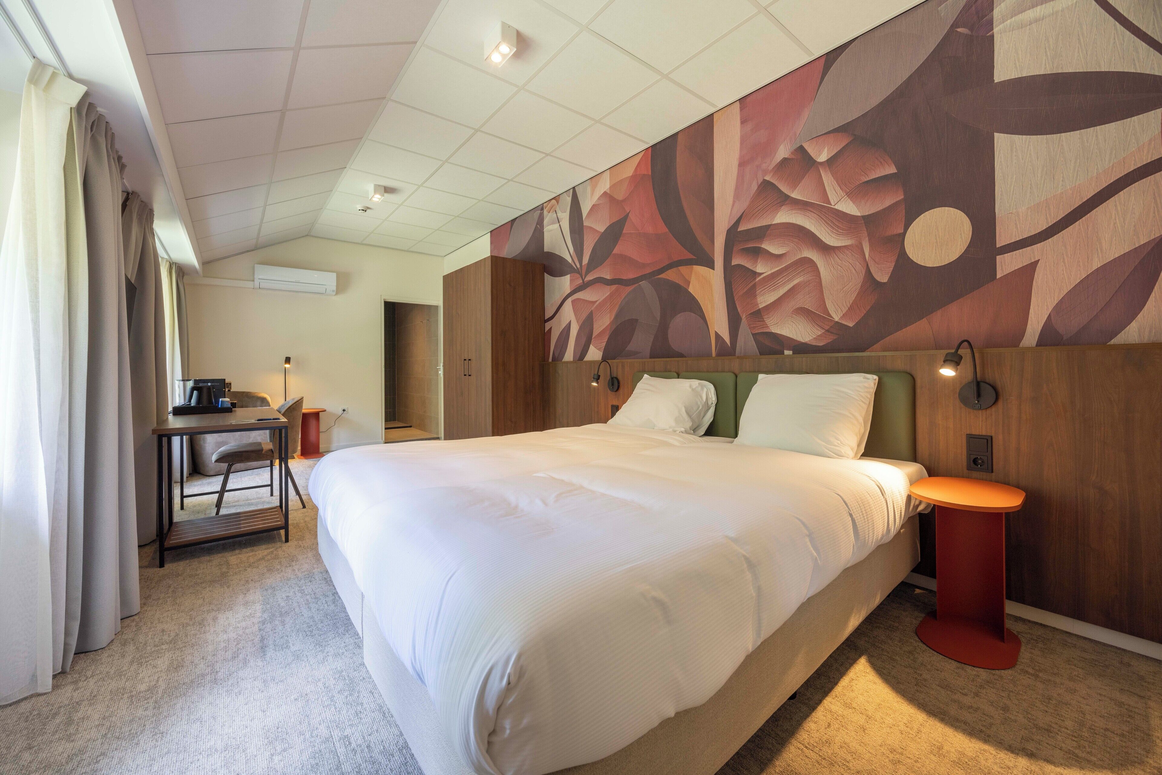 A hotel room with a large bed, a desk with a chair, a small table, and a wall with a floral mural.