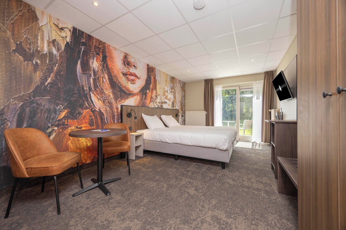 Habitación de hotel con cama, una silla, una mesita, televisor y un cuadro abstracto grande en la pared.