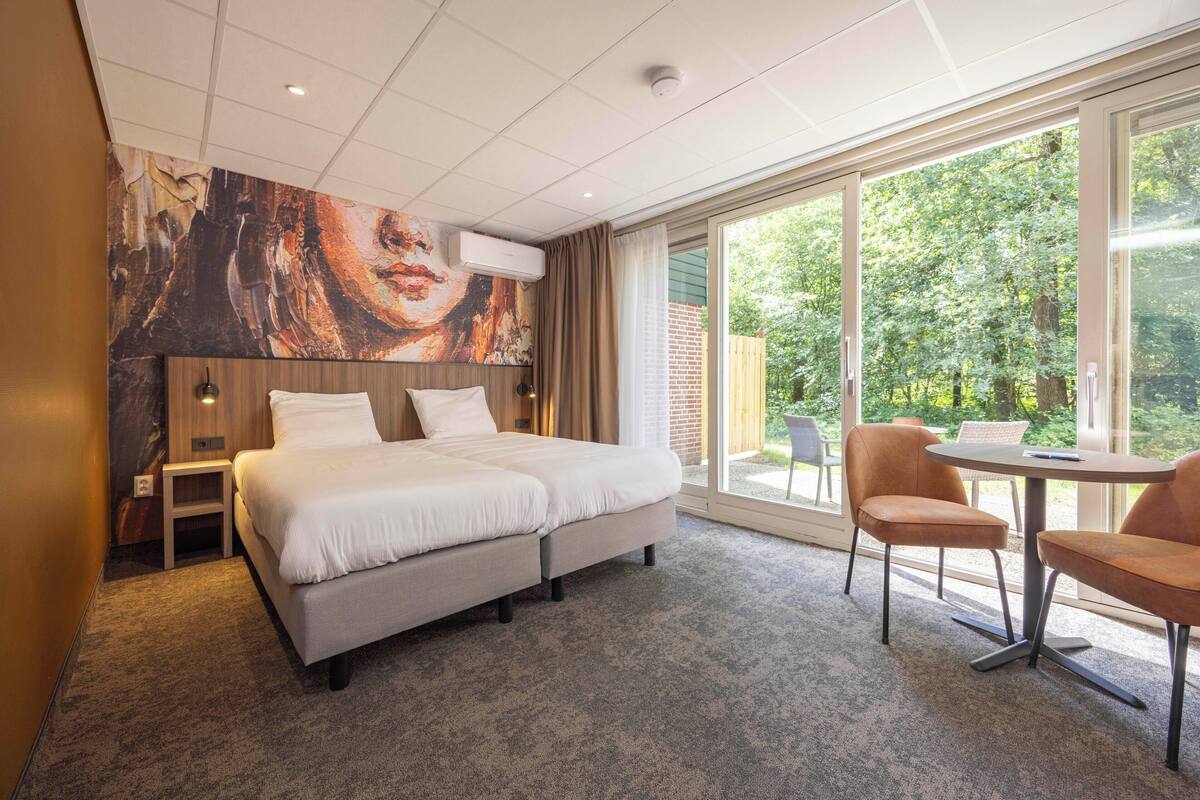A hotel room with a bed, a small table, a chair, and a large window offering a view of greenery.