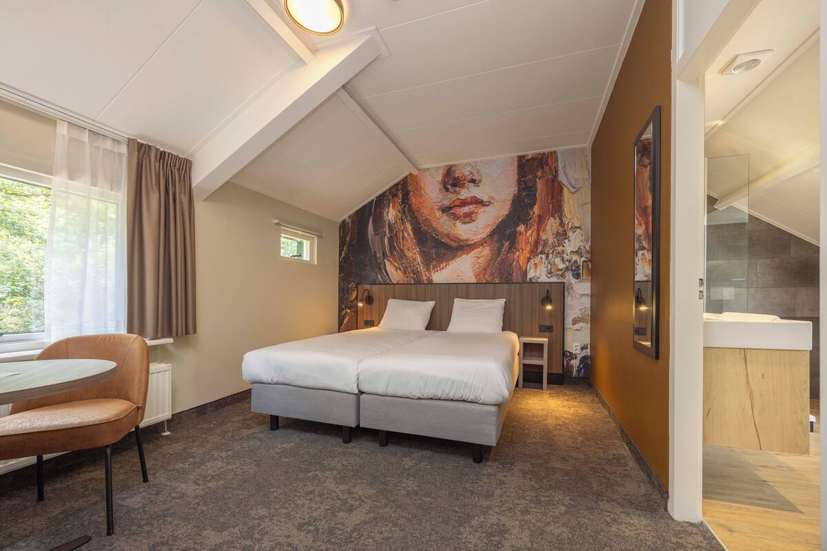 A hotel room with a bed, a desk, a chair, a window with curtains, and a painting on the wall.