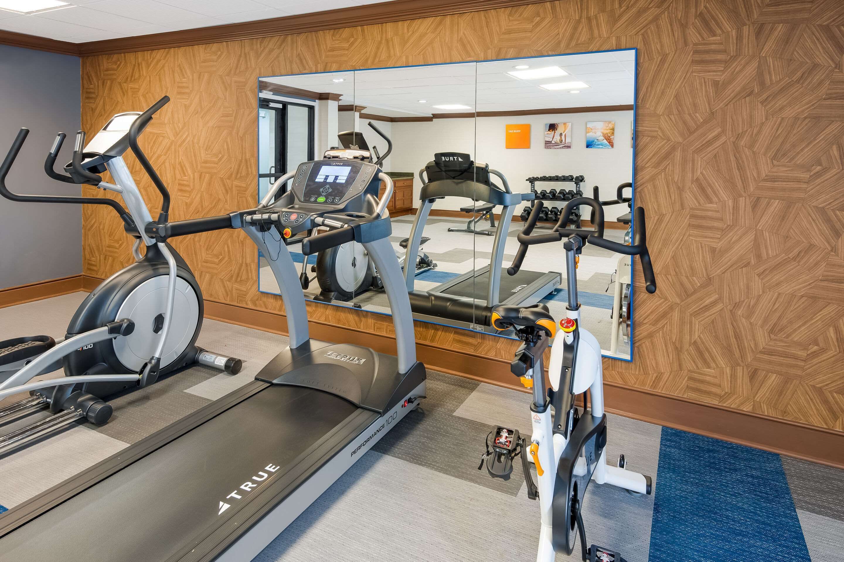 A gym with a treadmill, elliptical, and stationary bike, a mirror reflecting the room, and exercise equipment.