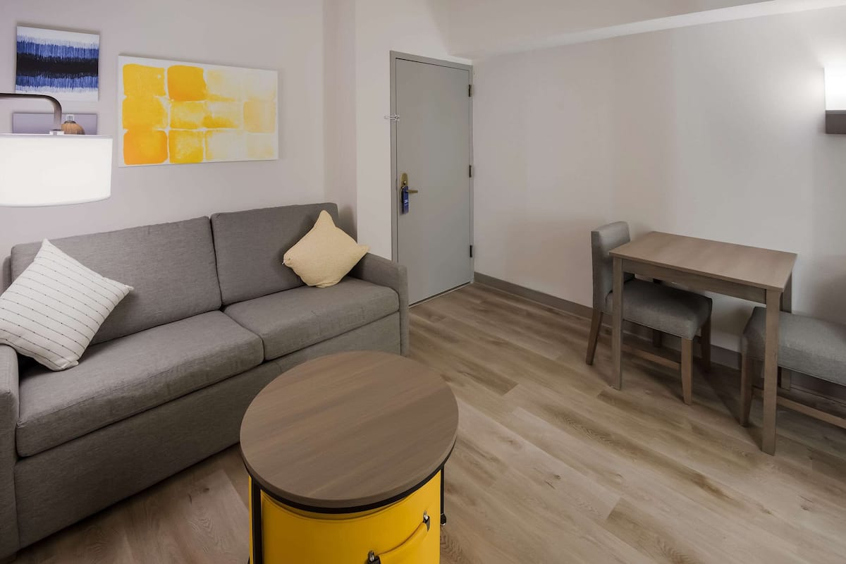 Una sala de estar moderna con un sofá gris, un tambor amarillo redondo, una mesa de madera con dos sillas y una puerta con manija.