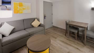 Un soggiorno moderno con un divano grigio, un tamburo giallo rotondo, un tavolo in legno con due sedie e una porta con maniglia.