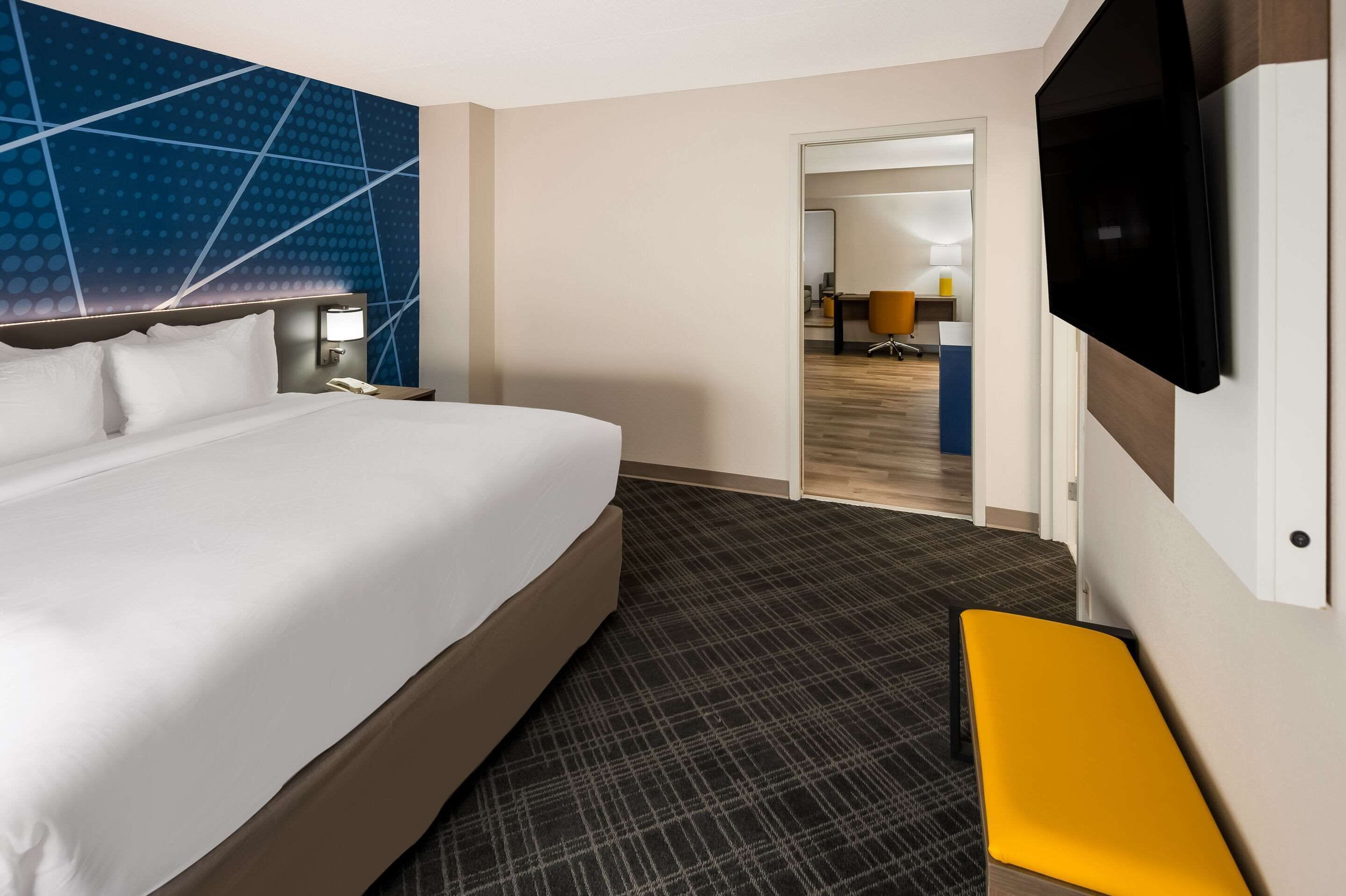 Habitación de hotel con una cama grande, un televisor de pantalla plana en la pared, un escritorio pequeño con lámpara y vista al interior a través de una puerta abierta.