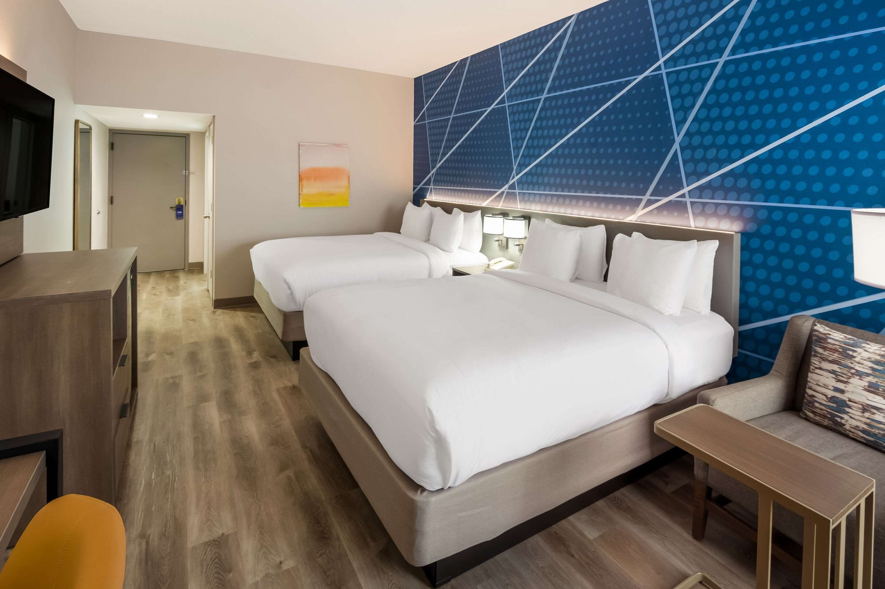 A hotel room with two beds, a desk, a TV, and a wall with a geometric pattern.