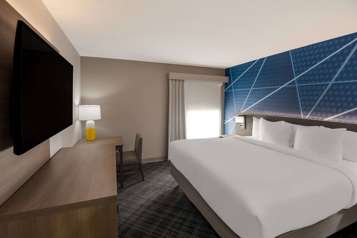 Habitación de hotel con una cama grande, un escritorio con lámpara, un televisor de pantalla plana y un ventanal con cortinas.