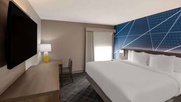 Habitación de hotel con una cama grande, un escritorio con lámpara, un televisor de pantalla plana y un ventanal con cortinas.
