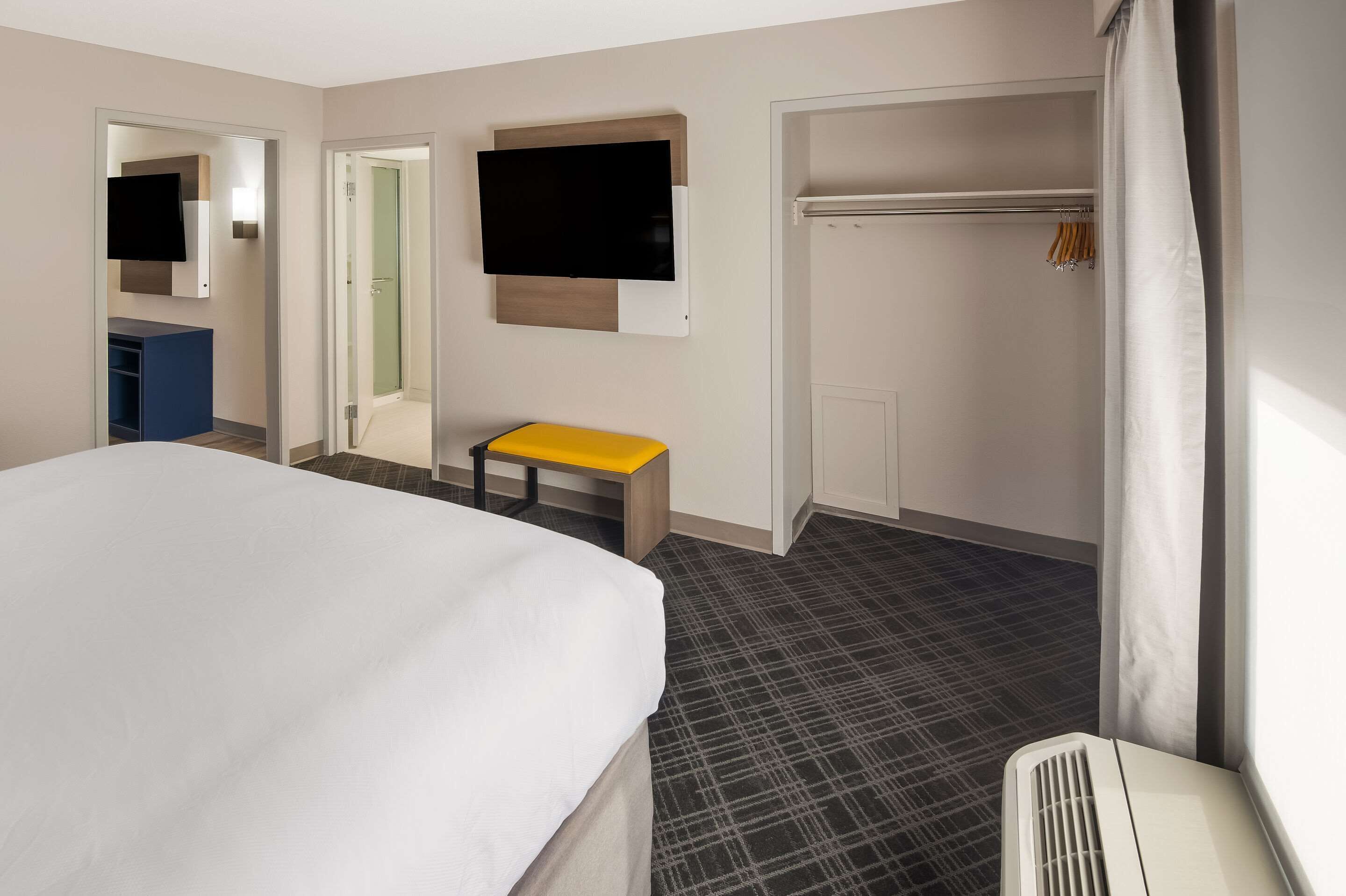 Habitación de hotel con cama, una mesita, televisor montado en la pared, armario y baño visible a través de una puerta abierta.