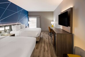 A hotel room with two beds, a desk with a chair, a flat-screen TV mounted on the wall, and a window with curtains.