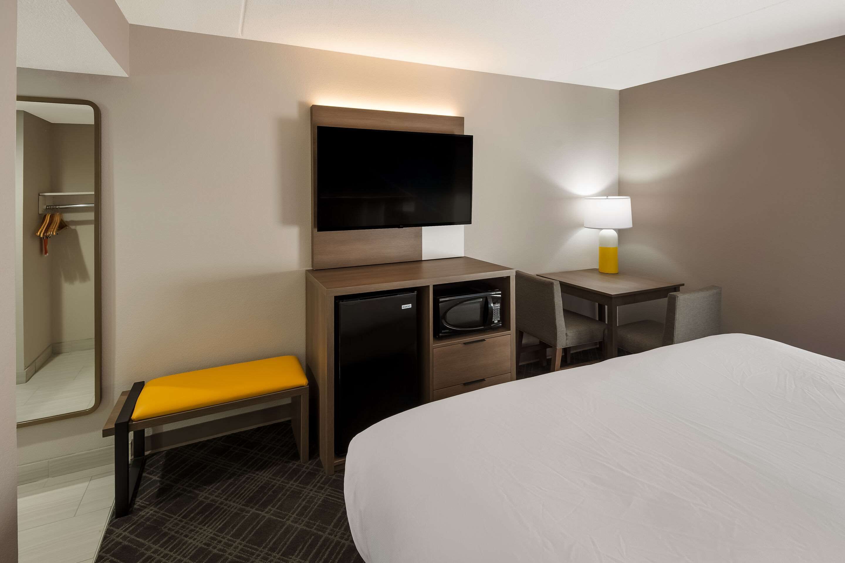 A hotel room with a bed, a desk with a lamp, a TV mounted on the wall, a microwave, a small refrigerator, and a bench.
