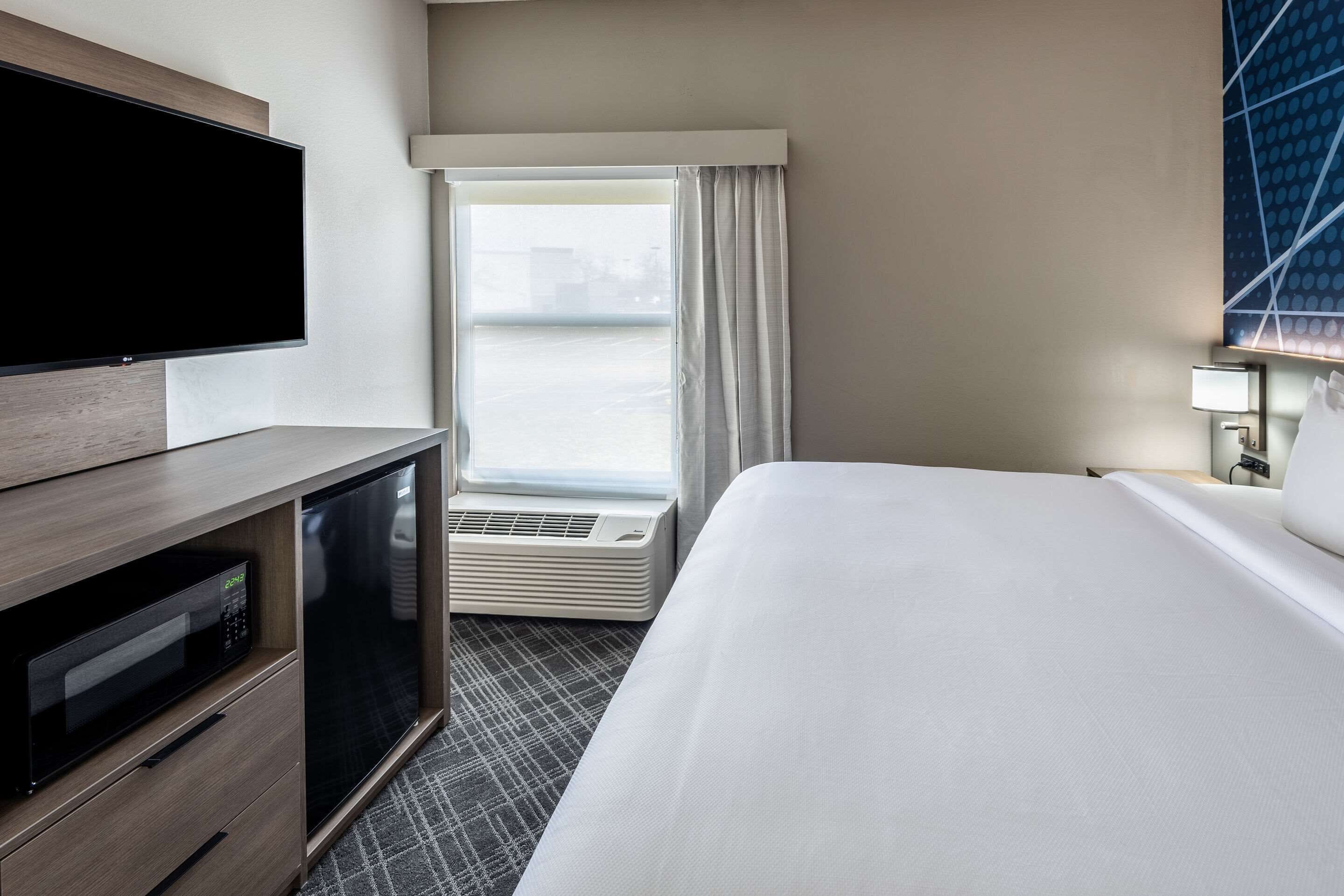 Une chambre d’hôtel équipée d’une télévision à écran plat, d’un mini-réfrigérateur, d’un four à micro-ondes, d’un grand lit, d’une fenêtre avec des rideaux et d’une lampe fixée au mur.