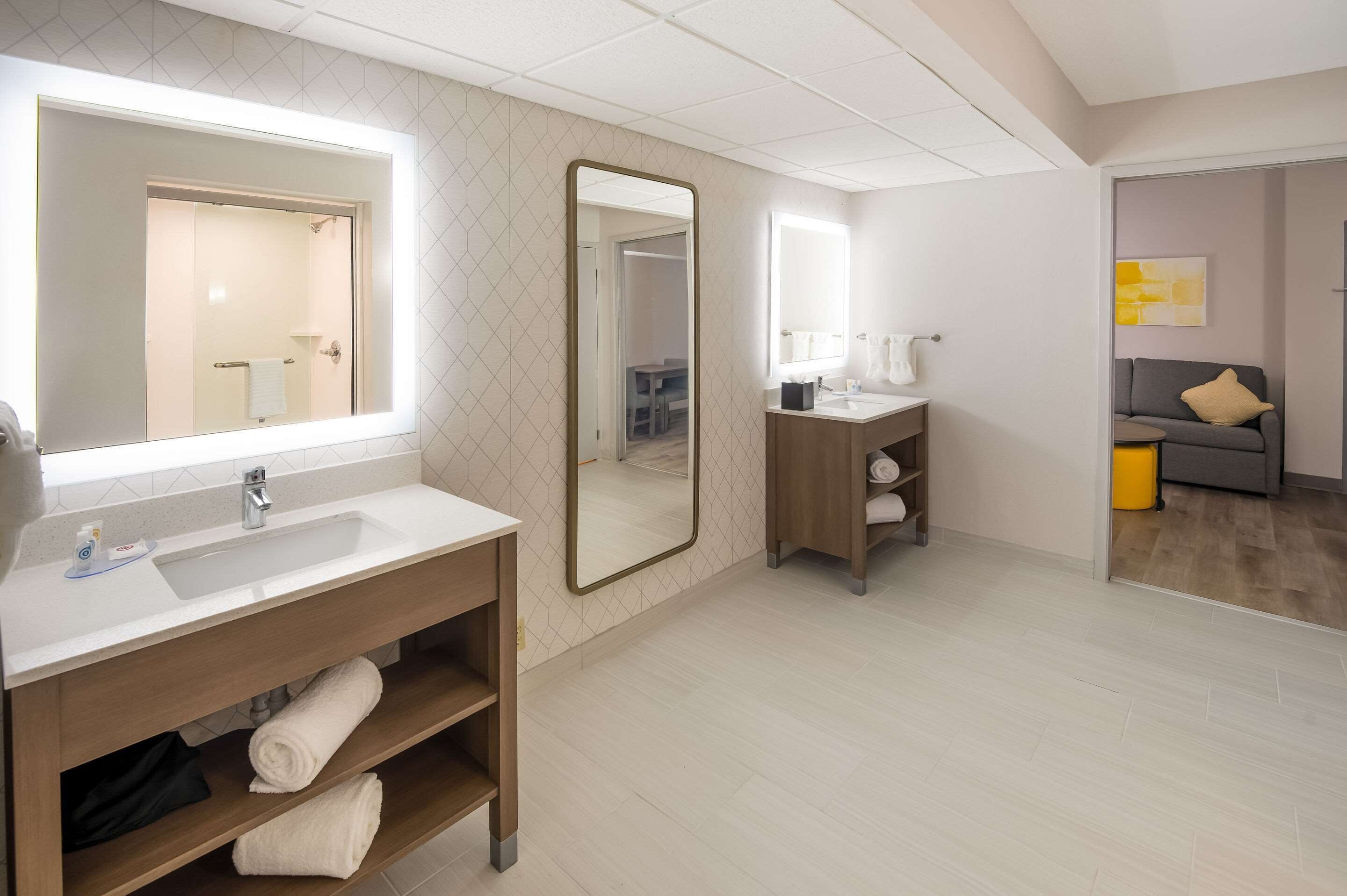 A modern bathroom with a large mirror, a sink, and a vanity. There is a towel rack and a shelf with towels.