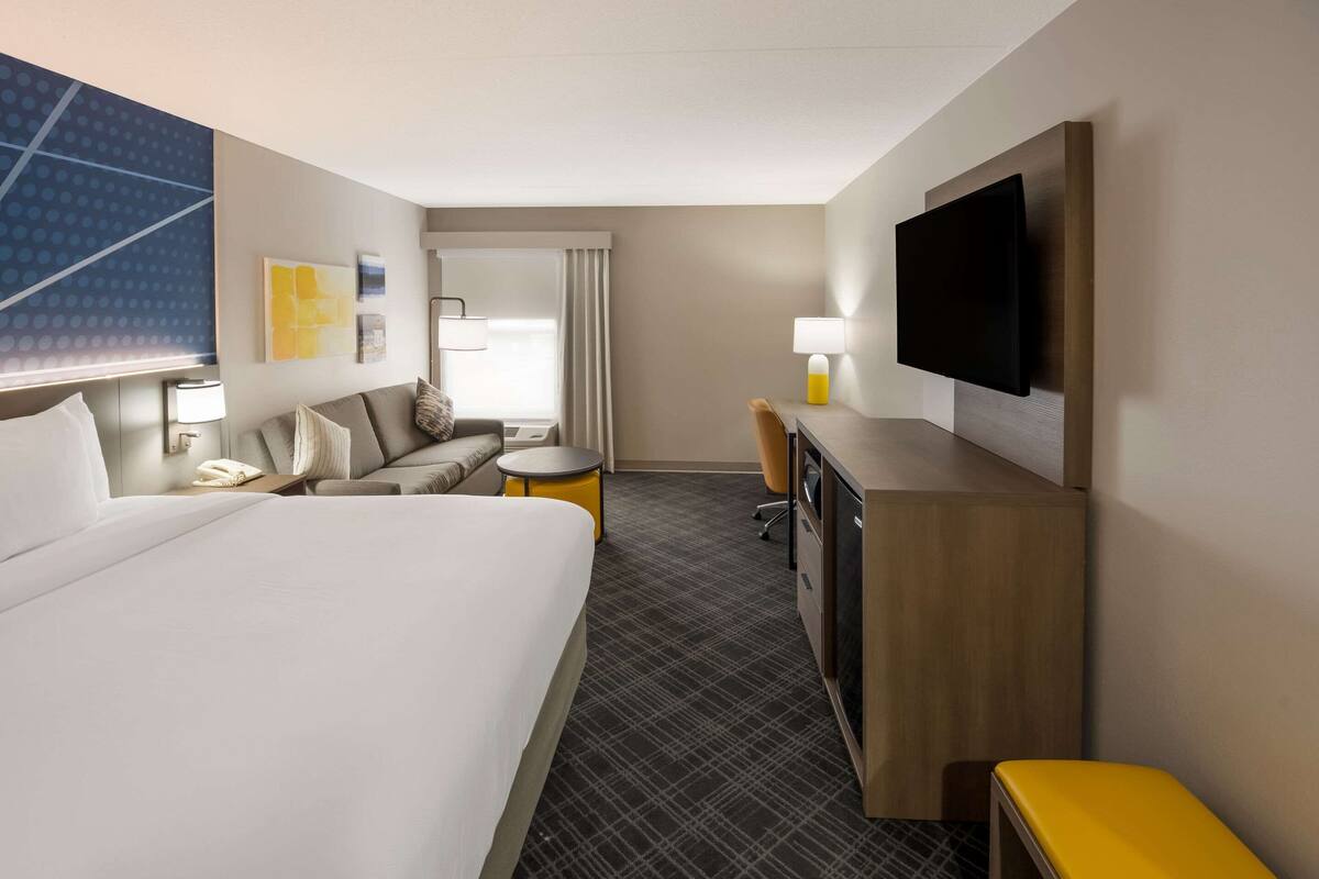 Habitación de hotel con una cama grande, un escritorio, una silla, un televisor montado en la pared y una mesita de noche con lámpara.