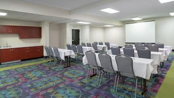 A conference room with tables, chairs, a projector screen, and a kitchen area.