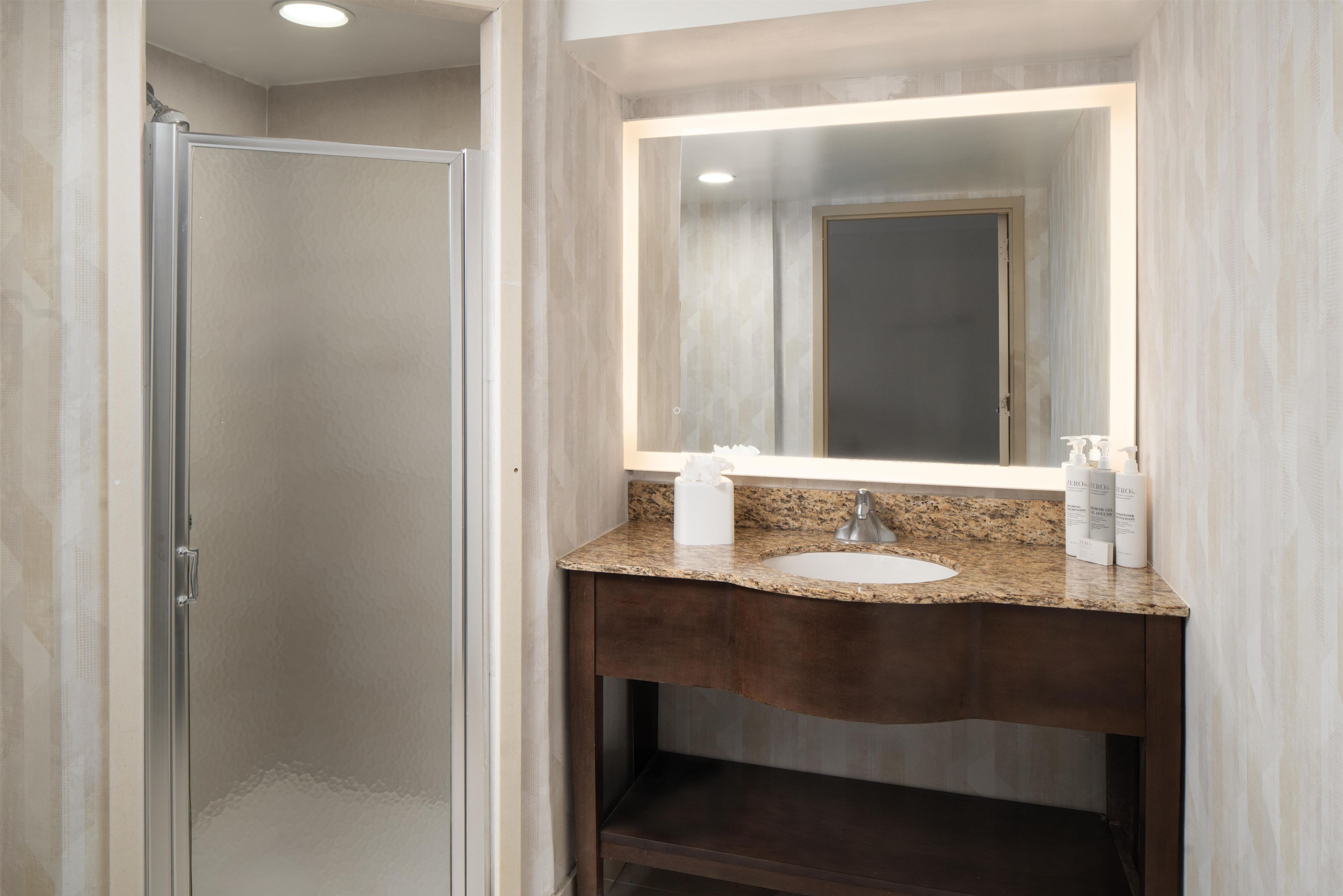 A bathroom with a glass shower enclosure, a granite countertop sink, and a mirror with built-in lighting.