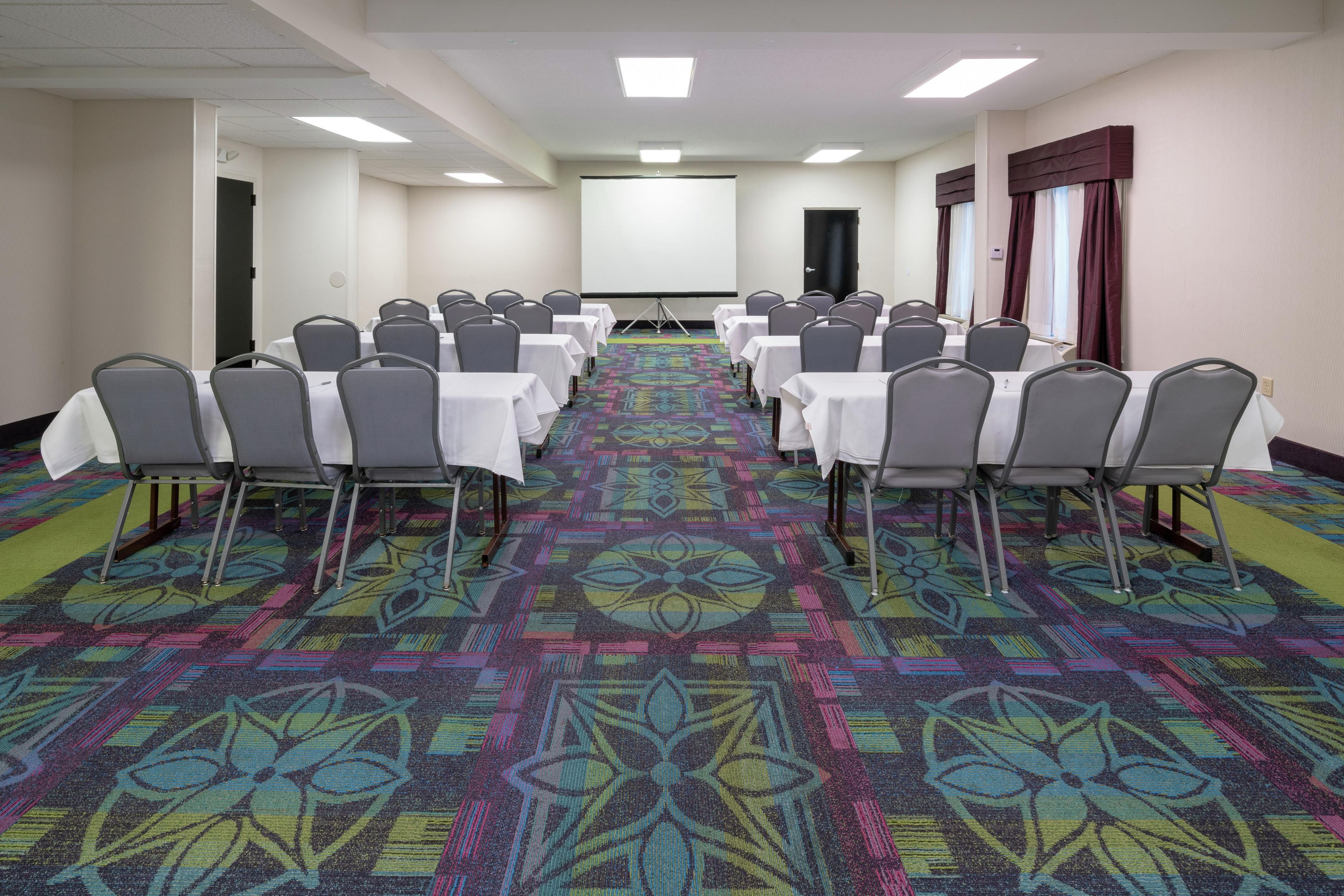 A conference room with rows of tables and chairs, a projector screen, and a carpeted floor.