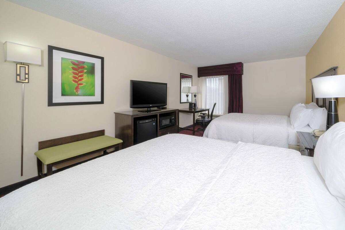 A hotel room with two beds, a television, a desk, a bench, and a framed picture on the wall.
