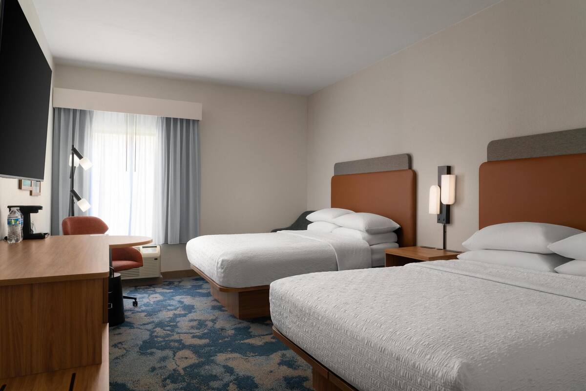 A hotel room with two beds, a desk, a chair, a TV, and a window with curtains.