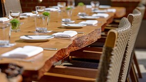 A long wooden dining table set with plates, glasses, and cutlery, surrounded by wicker chairs.
