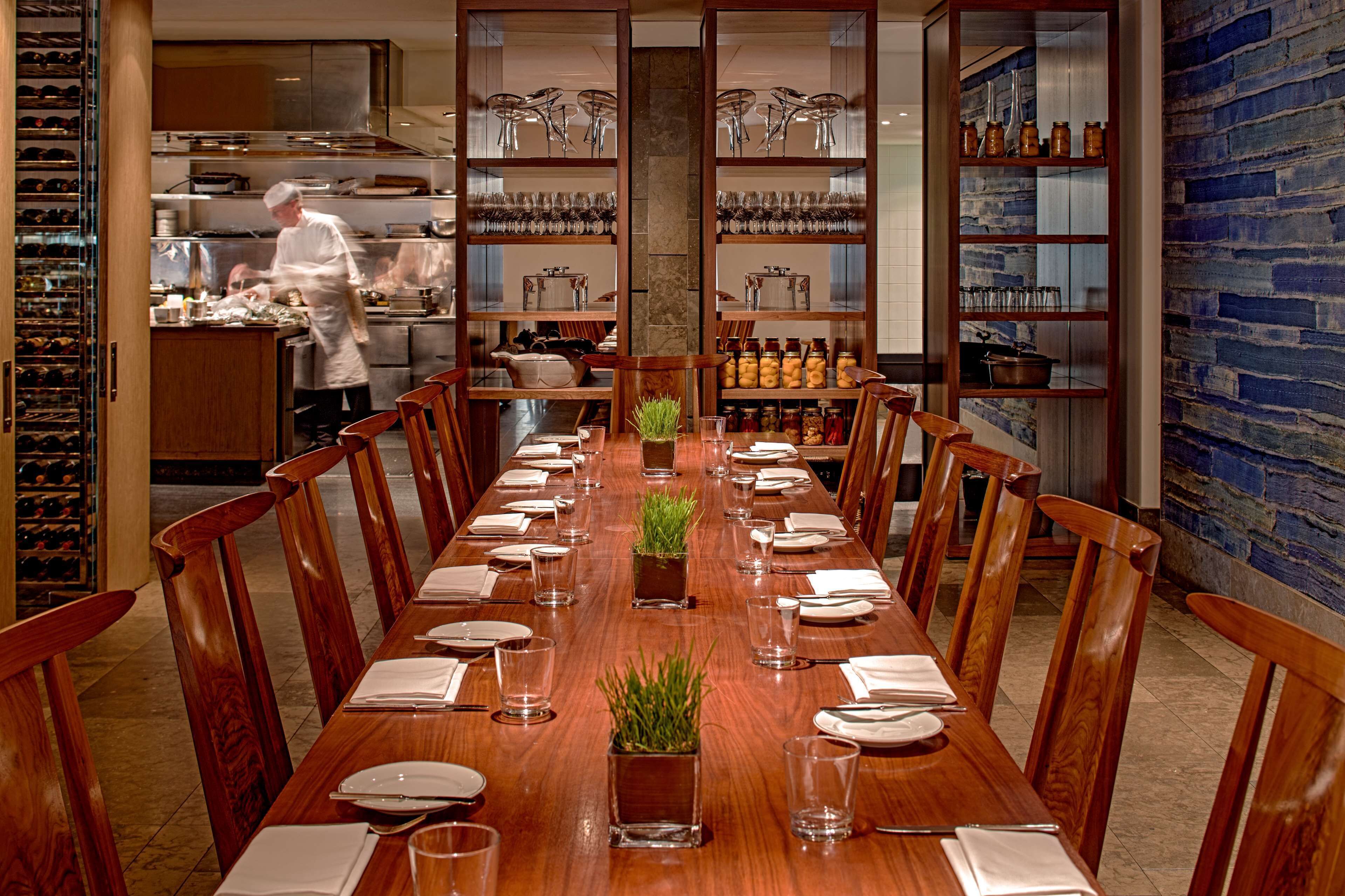 A dining area with a long wooden table set for a meal, flanked by wooden chairs. In the background, there is a kitchen area with a chef preparing food.