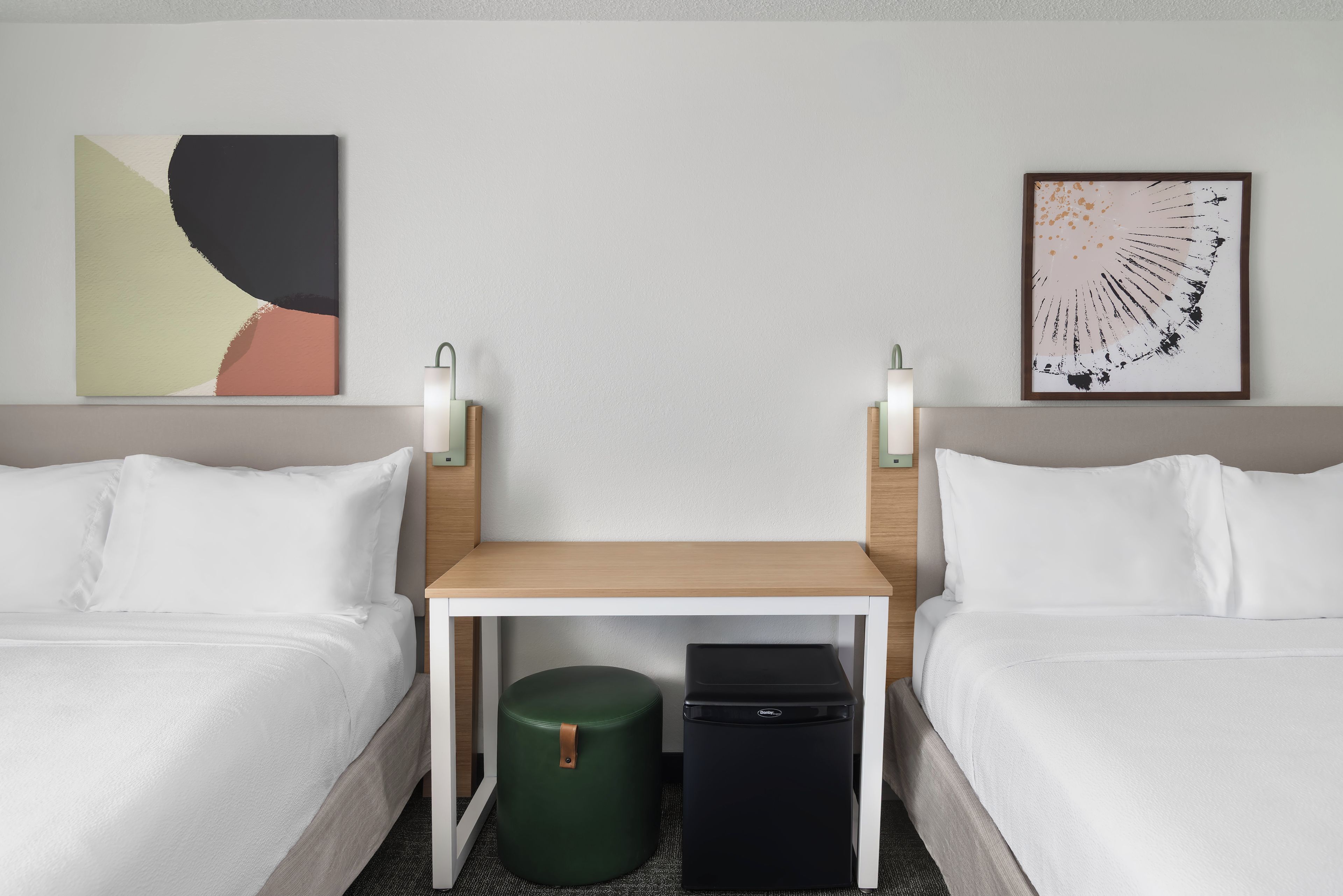 Habitación de hotel con dos camas, una mesita pequeña, un taburete verde y un mini bar negro.
