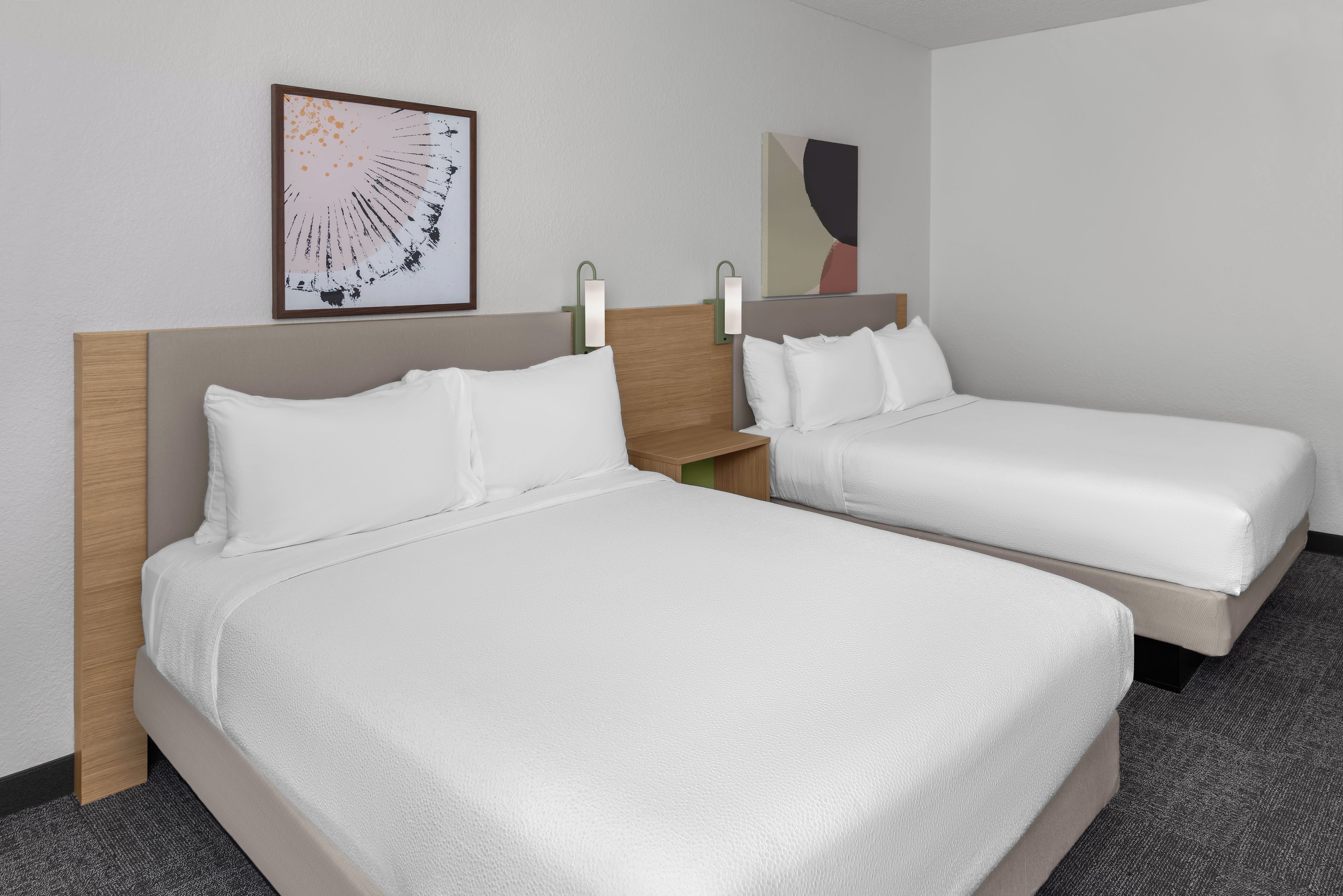 A hotel room with two beds, a headboard, a nightstand, a wall-mounted lamp, and two pieces of wall art.