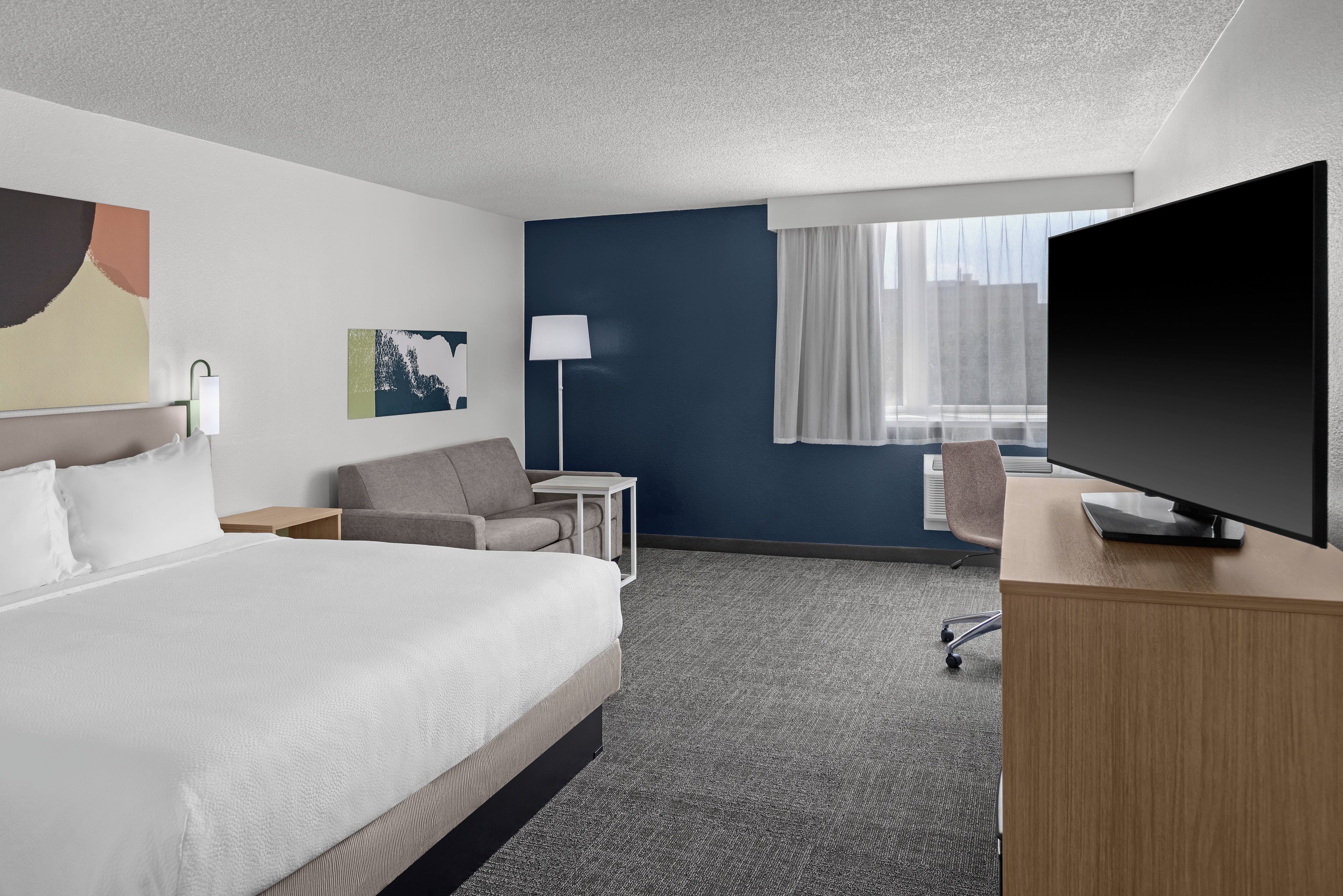 A hotel room with a bed, a desk, a chair, a TV, a lamp, and a window with curtains.