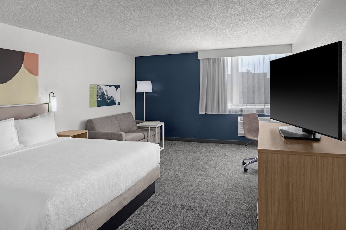 A hotel room with a bed, a desk, a chair, a TV, a lamp, and a window with curtains.