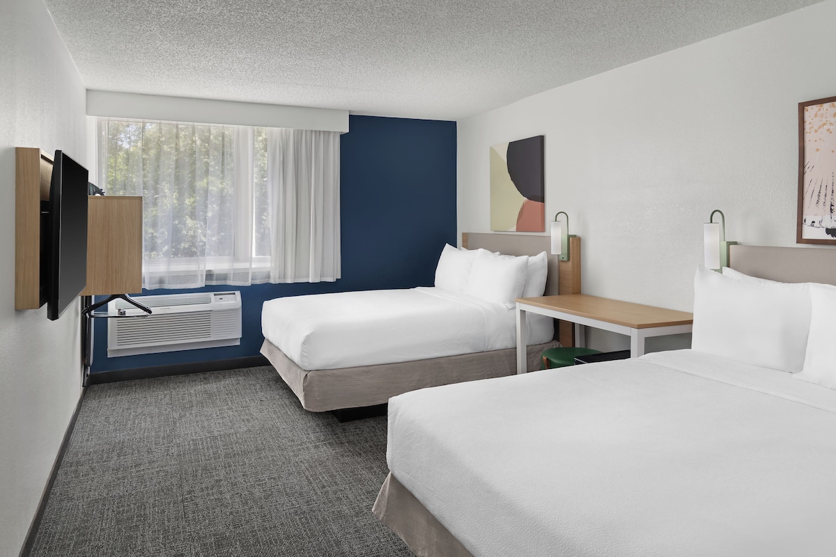 A hotel room with two beds, a TV, a desk, a lamp, and a window with curtains.