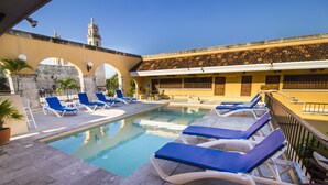 Un espace piscine avec des chaises longues, un mur en pierre et un bâtiment doté d’arches et d’un clocher en arrière-plan.