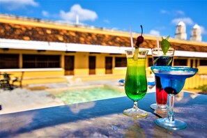 A poolside bar with two cocktails, a clear pool, and a building with a yellow facade in the background.