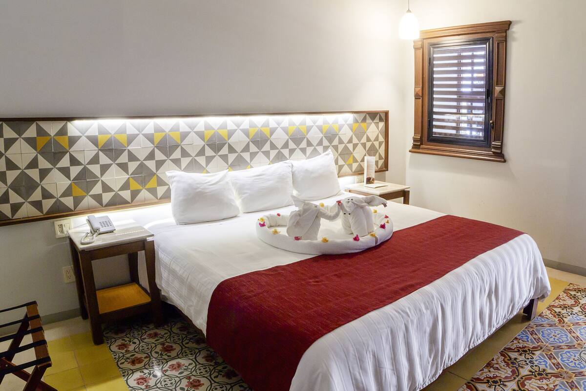 A neatly made bed with a red blanket, a bedside table with a phone, and a wall with geometric patterned tiles.