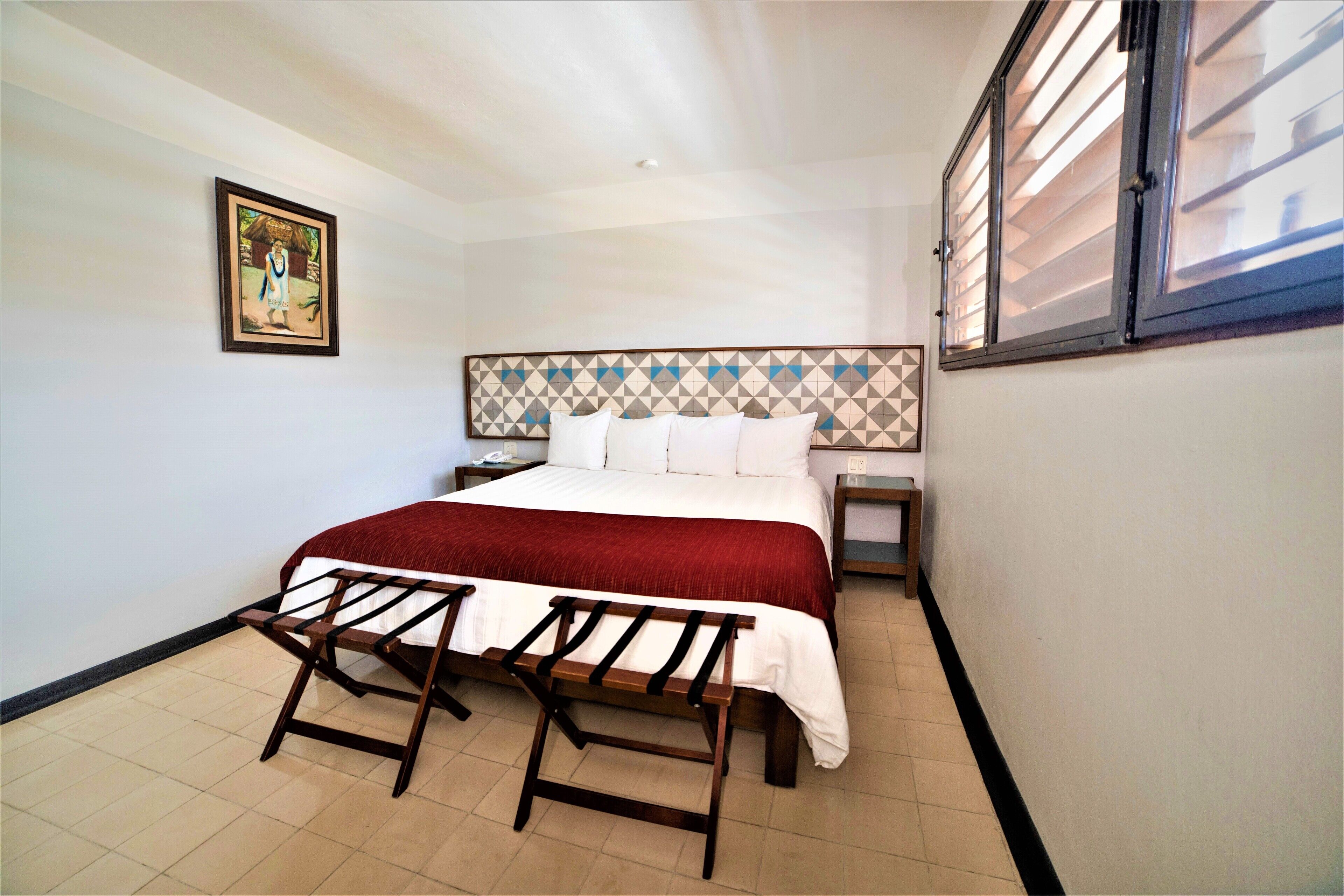 A bedroom with a bed, a nightstand, a bench, a wall-mounted picture, and a window with blinds.