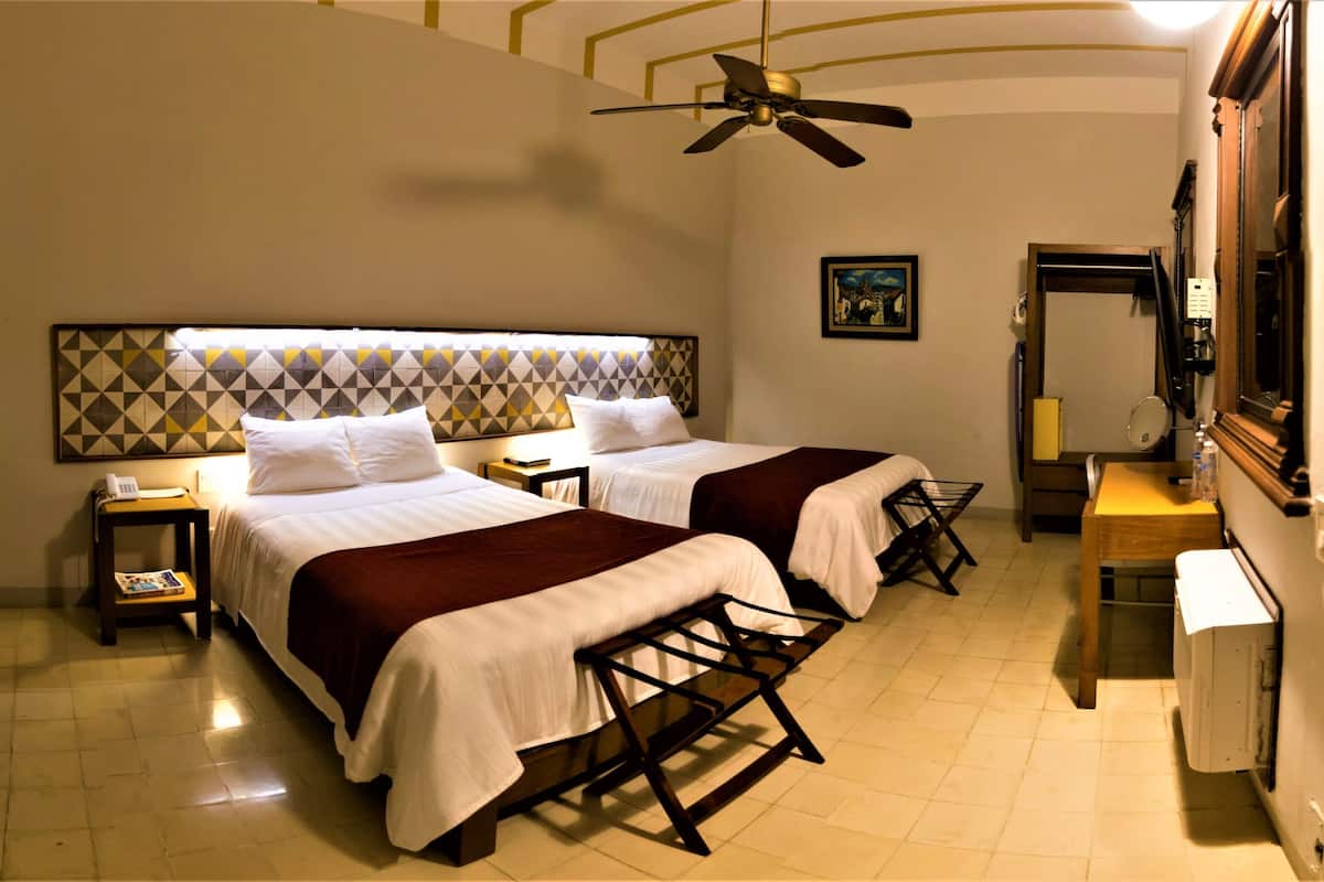 A hotel room with two beds, a ceiling fan, a desk, a chair, a mirror, and a radiator.