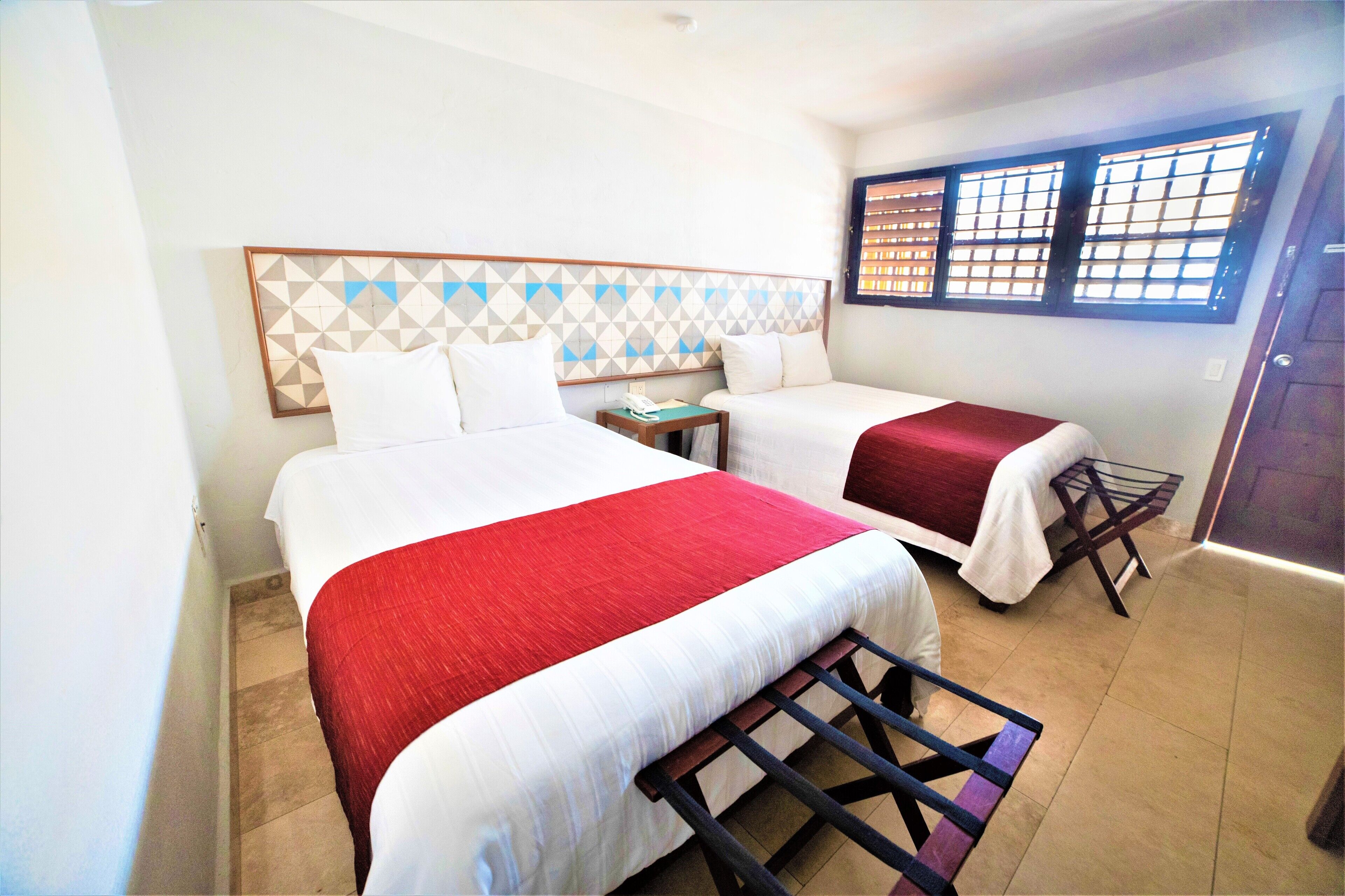 A hotel room with two beds, a headboard with a geometric pattern, a small table with a phone, a window with blinds, and a door.