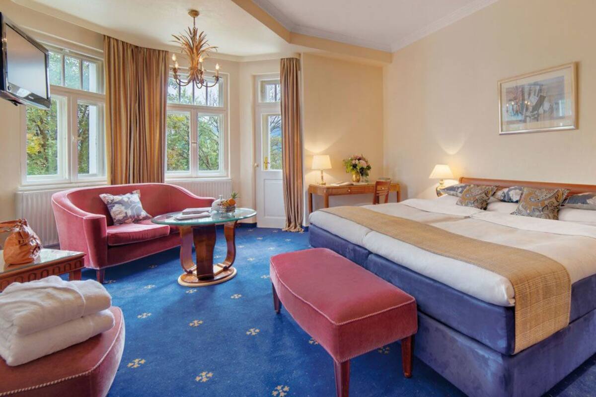 Een hotelkamer met een groot bed, een rode fauteuil, een klein tafeltje met een lamp en uitzicht op het omliggende landschap door grote ramen.