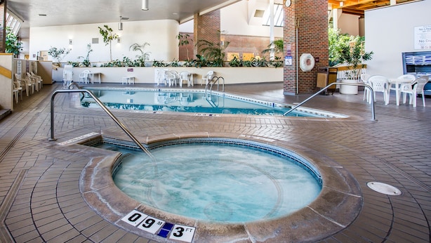 An indoor swimming pool with a hot tub, surrounded by white chairs and a brick wall.