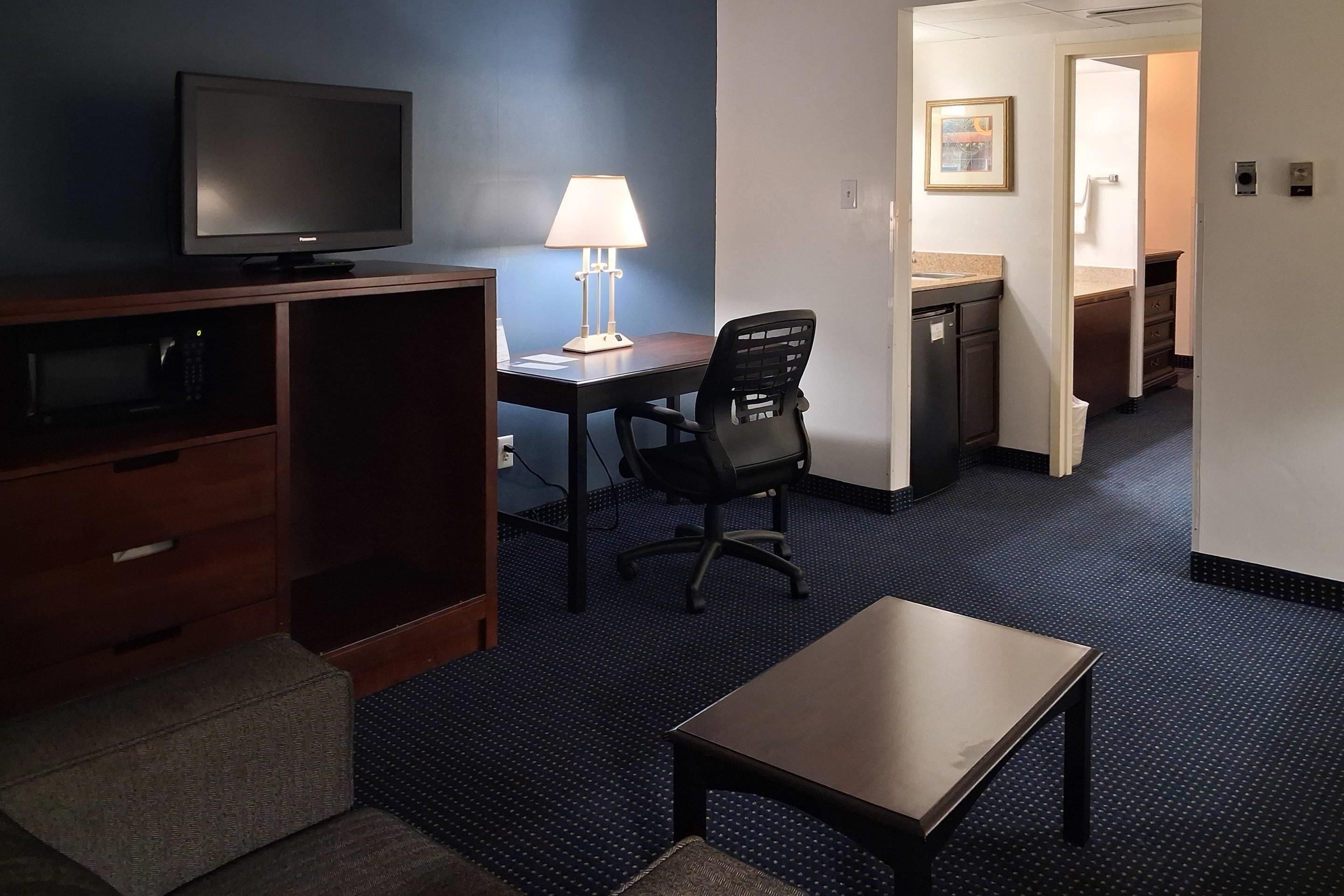 A hotel room with a TV, desk, chair, lamp, and a small table.