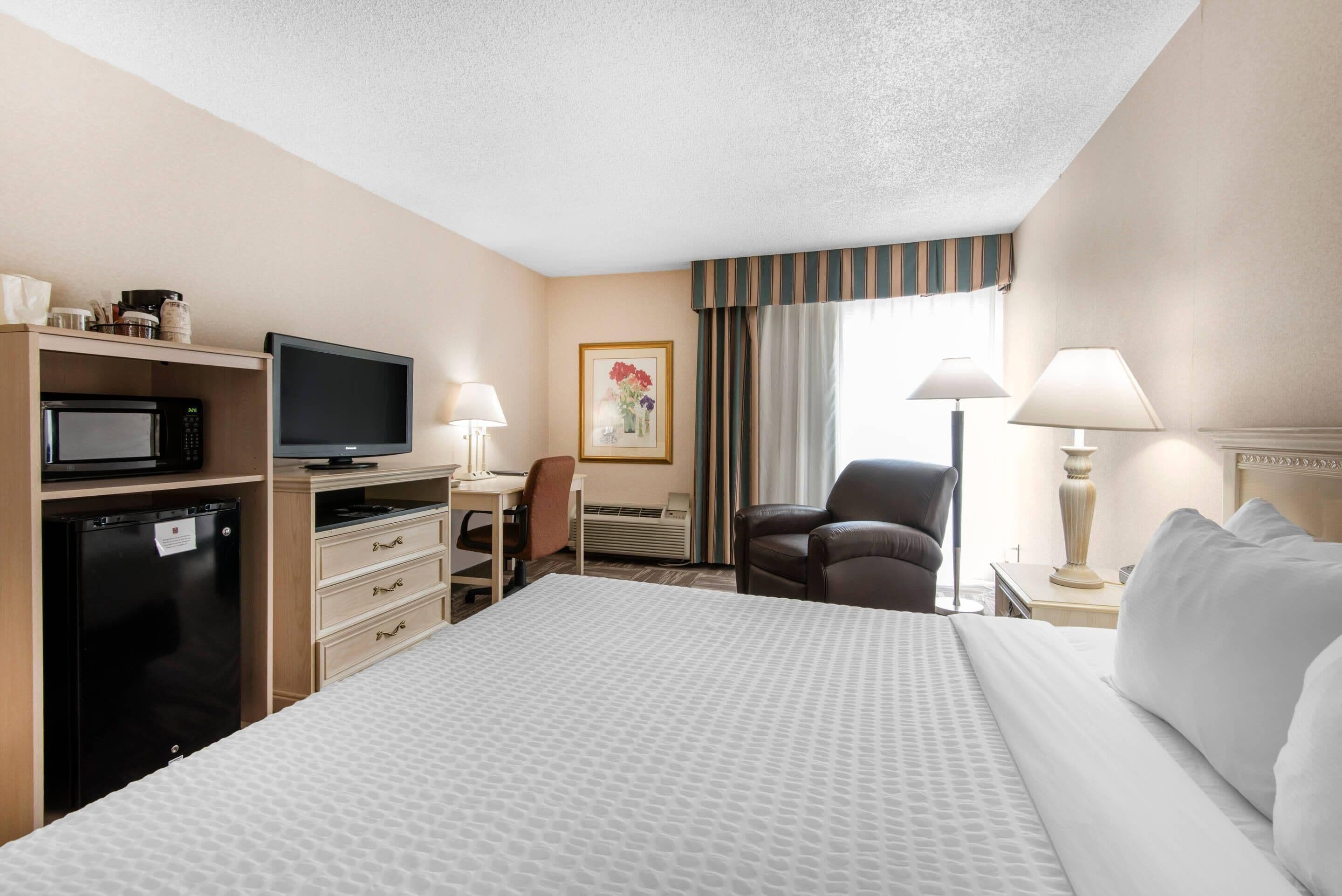 A hotel room with a bed, a desk with a computer, a television, a refrigerator, a chair, and a lamp.