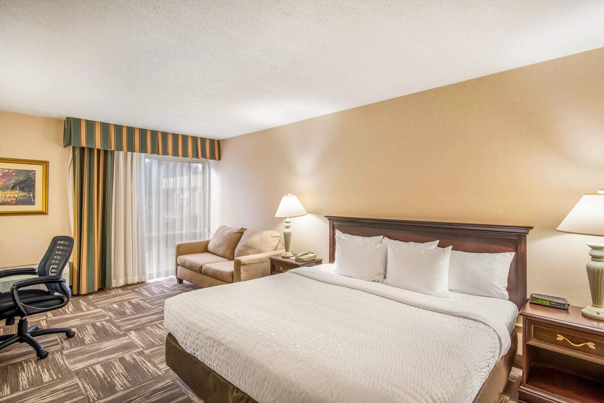 A hotel room with a large bed, a desk, a chair, a lamp, and a nightstand.