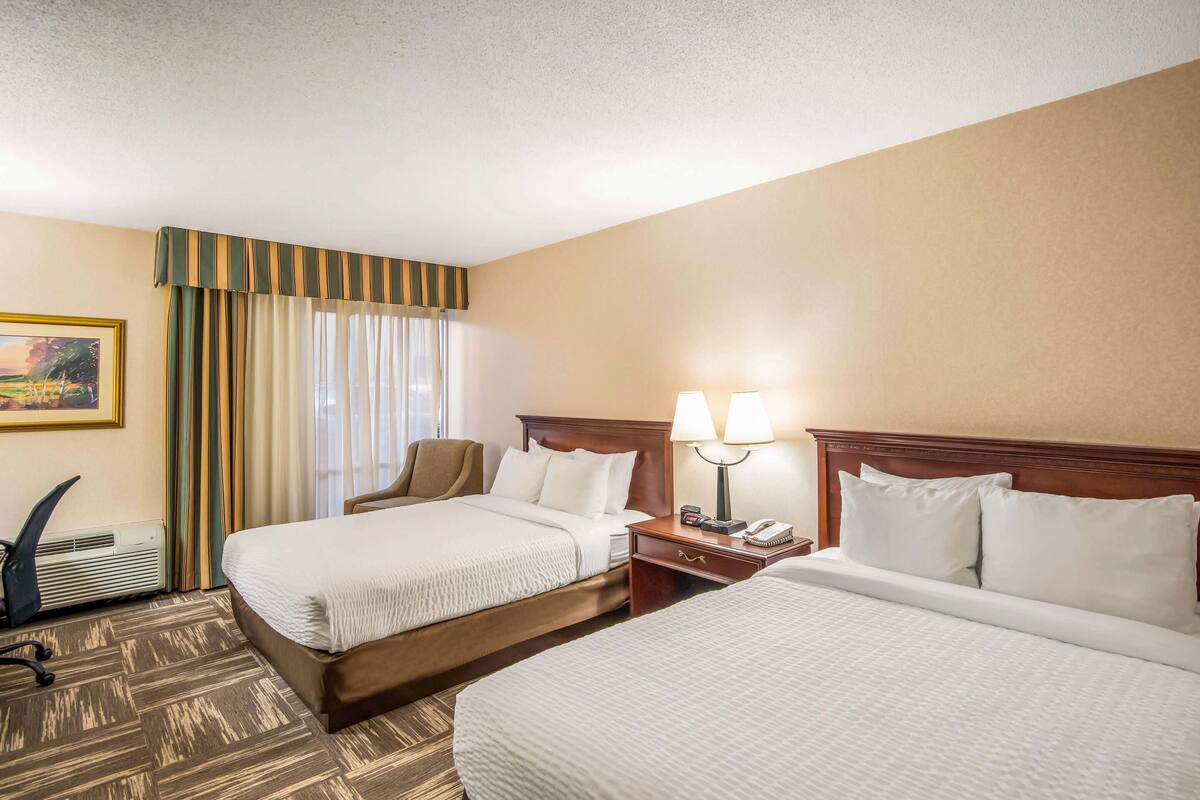 A hotel room with two beds, a desk with a lamp, a chair, a painting on the wall, and a window with curtains.