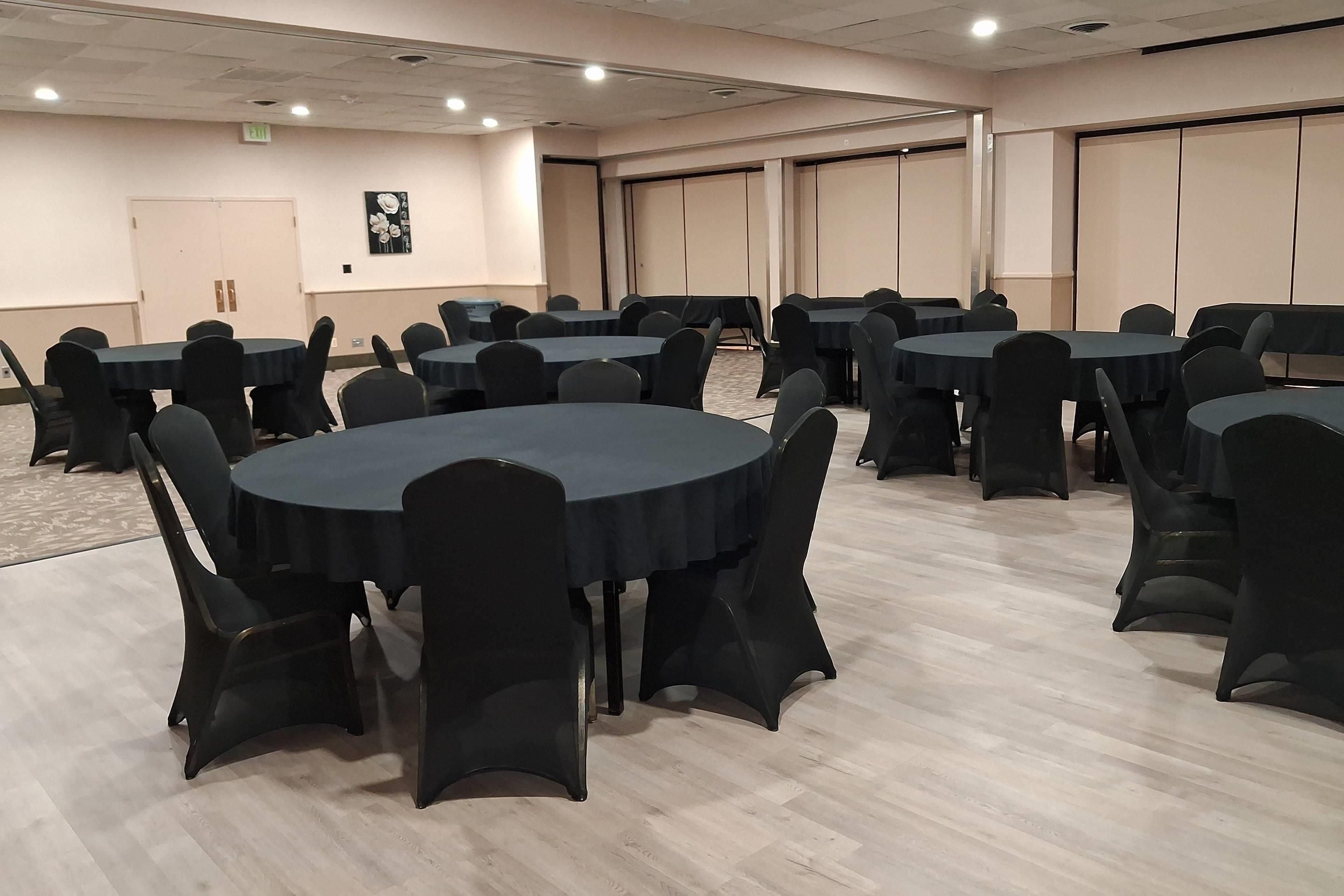 A conference room with multiple round tables and chairs, carpeted flooring, and a plain wall with a door and a framed picture.
