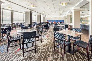 A spacious hotel dining area with multiple tables and chairs, large windows, and a kitchen area in the background. - Hilton Garden Inn San Francisco/Oakland Bay Bridge (Emeryville)
