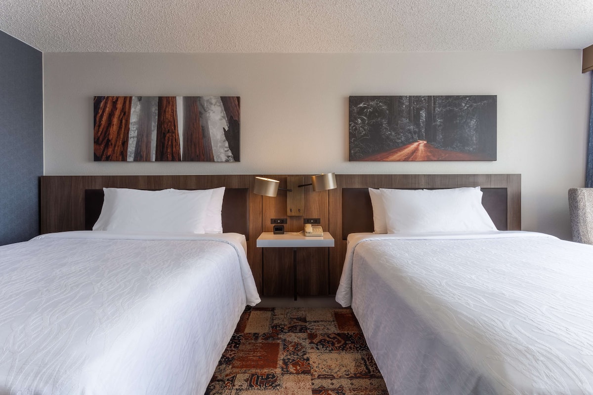 Een hotelkamer met twee bedden, een houten hoofdbord, een klein tafeltje met een lamp en kunstwerken aan de muur.