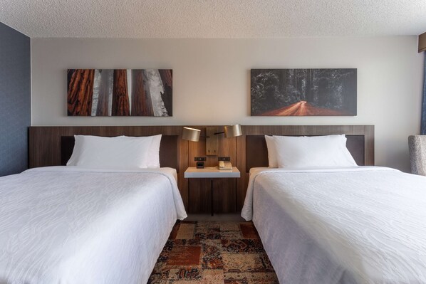 A hotel room with two beds, a wooden headboard, a small table with a lamp, and artwork on the walls.